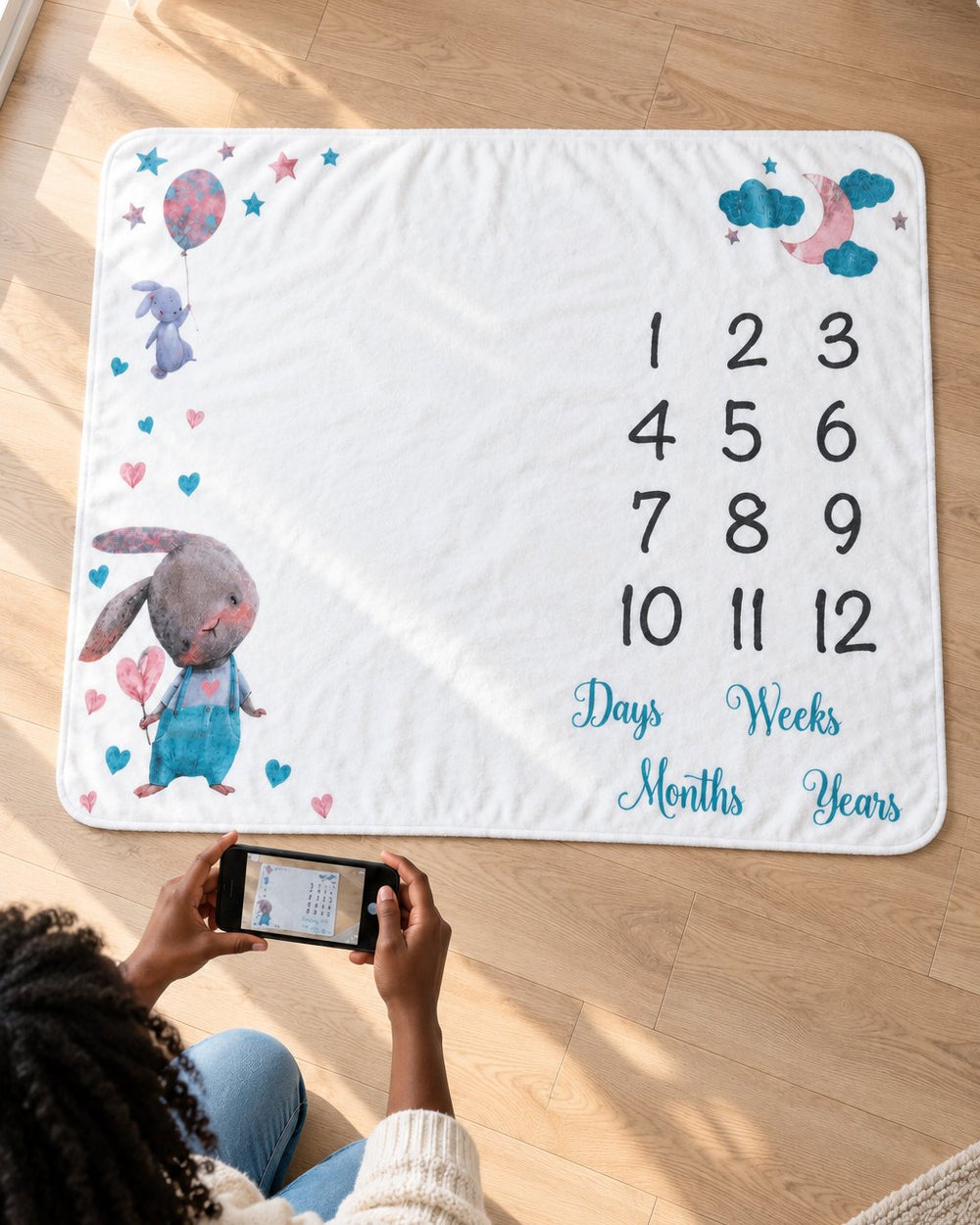 Milestone blanket spread on a wooden floor beside a parent kneeling with a phone