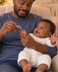 Dad hesitating with tiny nail clippers over his wriggling baby's hand in a nursery armchair
