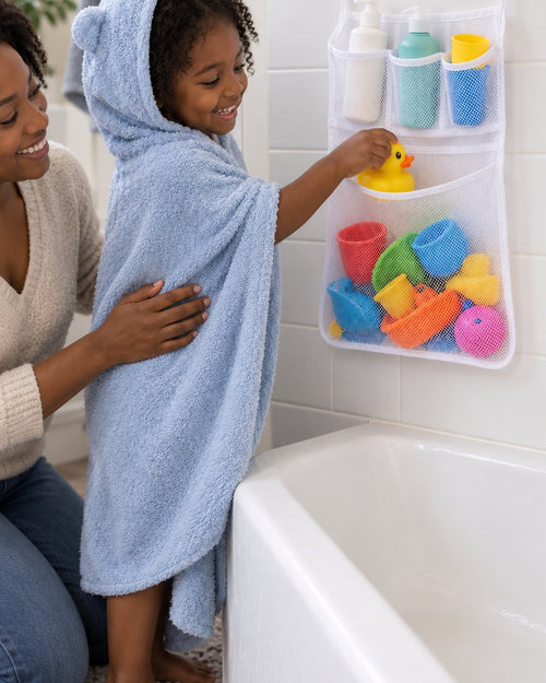 Child in a hooded towel dropping a rubber duck into the mesh net on the bathroom tile, a parent beside them