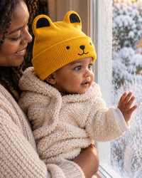 Parent holding a smiling baby in the bear beanie by a frosty window