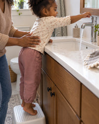 Small child on a step stool stretching to reach a bathroom tap, sleeve wet
