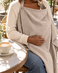 Mother at a cafe table wearing the oat muslin nursing cover, glancing down at her baby