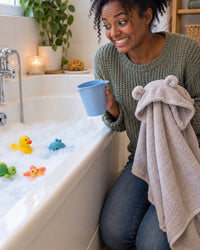 Parent kneeling by a bubble bath with a rinse cup and a hooded towel, bracing for hair-wash time