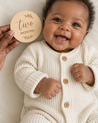 Awake baby on a flat play mat while a parent holds the 'two months' wooden disc beside them